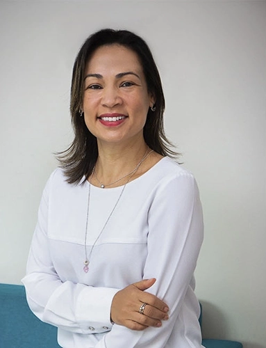 rejane em pé, com um braço cruzando o corpo e segurando no outro braço, sorrindo
