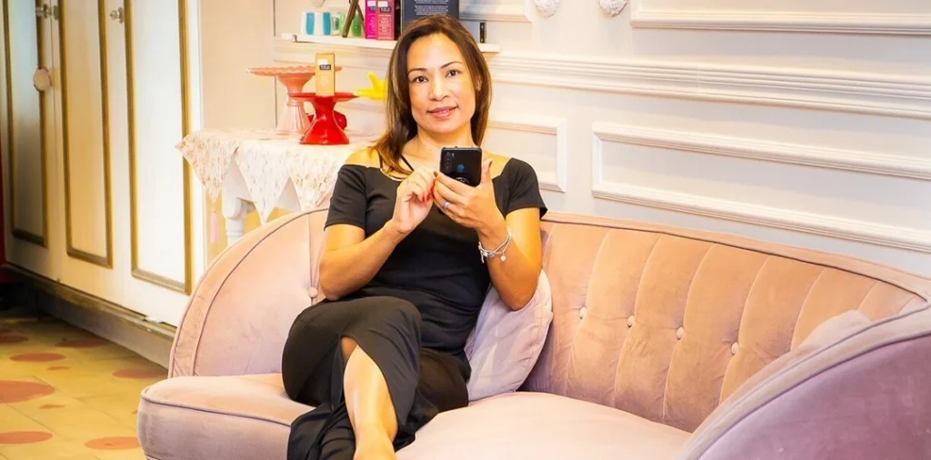 Rejane sentada em uma poltrona, sorrindo enquanto mexe no celular