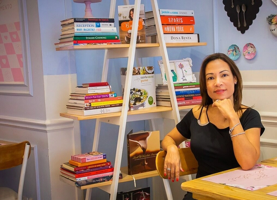 Rejane sentada em uma mesa, com a cabeça apoiada na mão, sorrindo