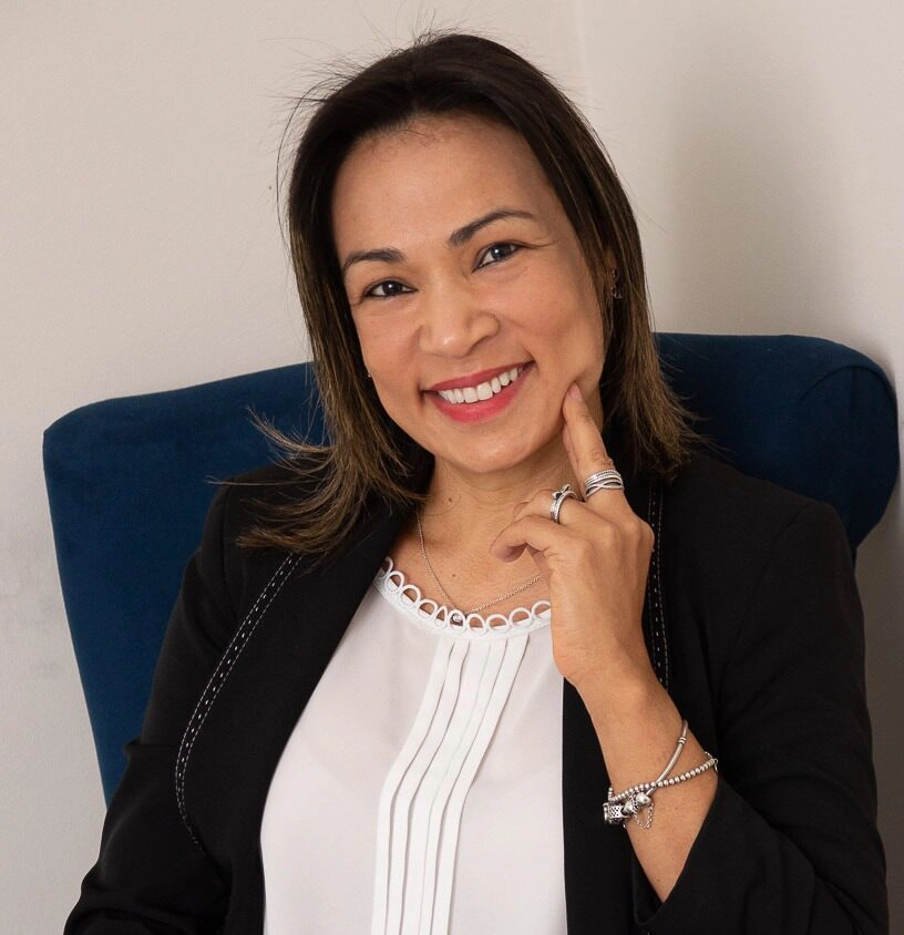 Rejane sentada em uma cadeira azul, com o rosto apoiado na mão, sorrindo